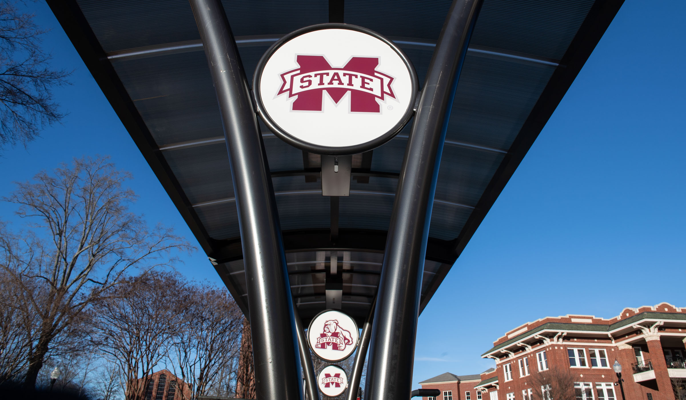 Three M-State logos on white circles decorate the center poles supporting the curved roof of a bike rack shelter, with blue sky, the Chapel, and the YMCA Building beyond.