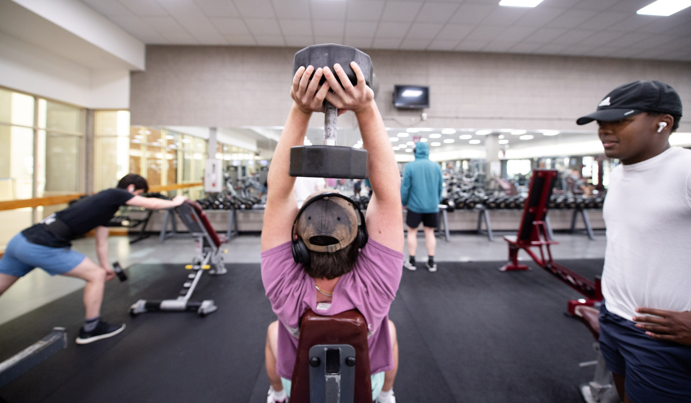 Sanderson weight room scene, with male student at center with back to viewer, lifting weight over head. Friend looks on from the right and other students lift in the background.