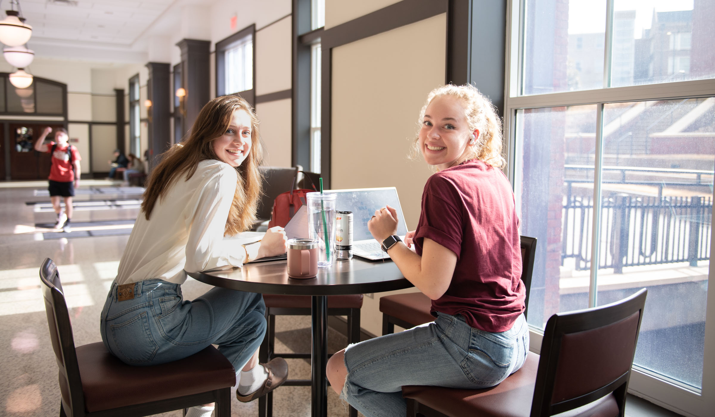 Two students sit in a sunbeam at an Old Main Academic Center between classes, smiling over their shoulders at the camera.