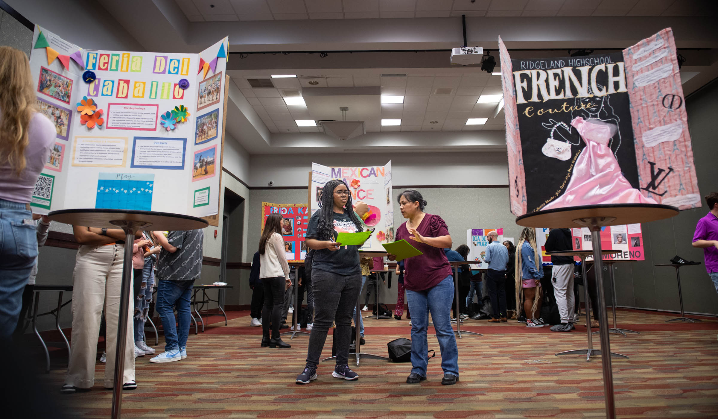 Surrounded by the bright colors of global culture posters on display, an MSU student judge talks with an CMLL Professor in the center.