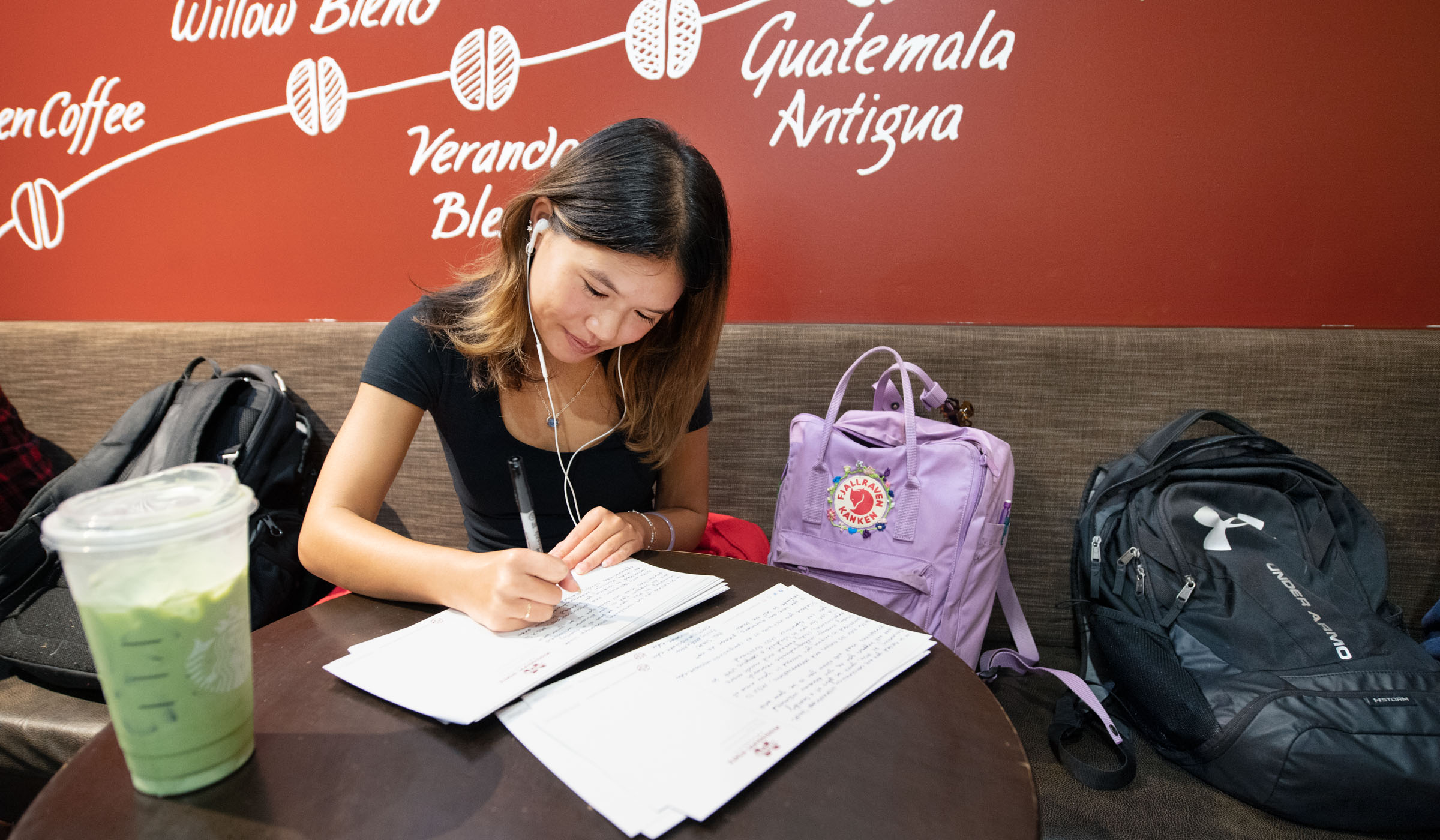 Orientation Leader Emily writes postcards to prospective MSU students at a Starbucks table, surrounded by backkpacks and with a green iced tea on hand.