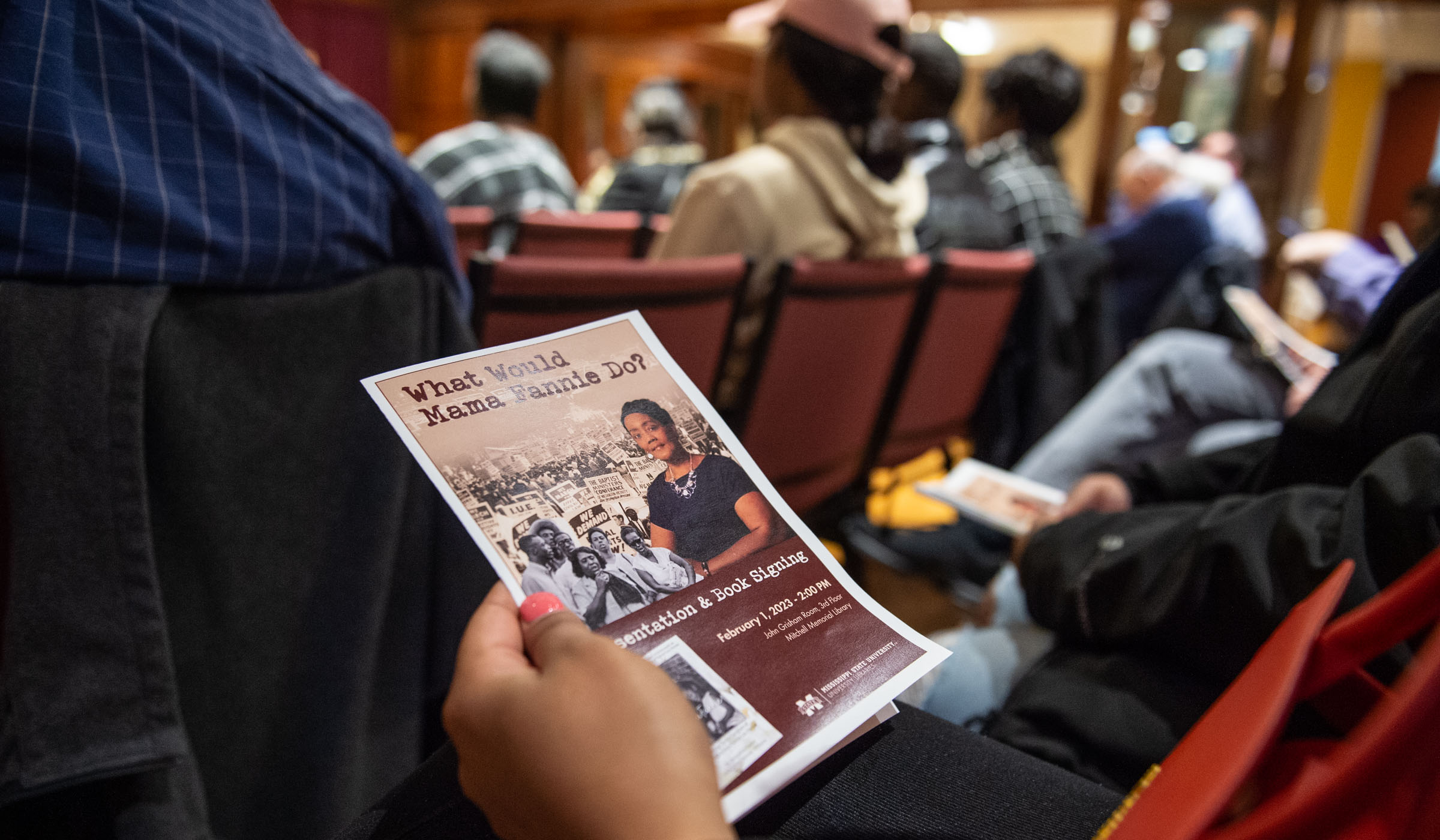 A &quot;What Would Mama Fannie Do?&quot; event program is held in the hand of an event attendee in the foreground, with the Grisham Room and other seated attendees out of focus beyond.