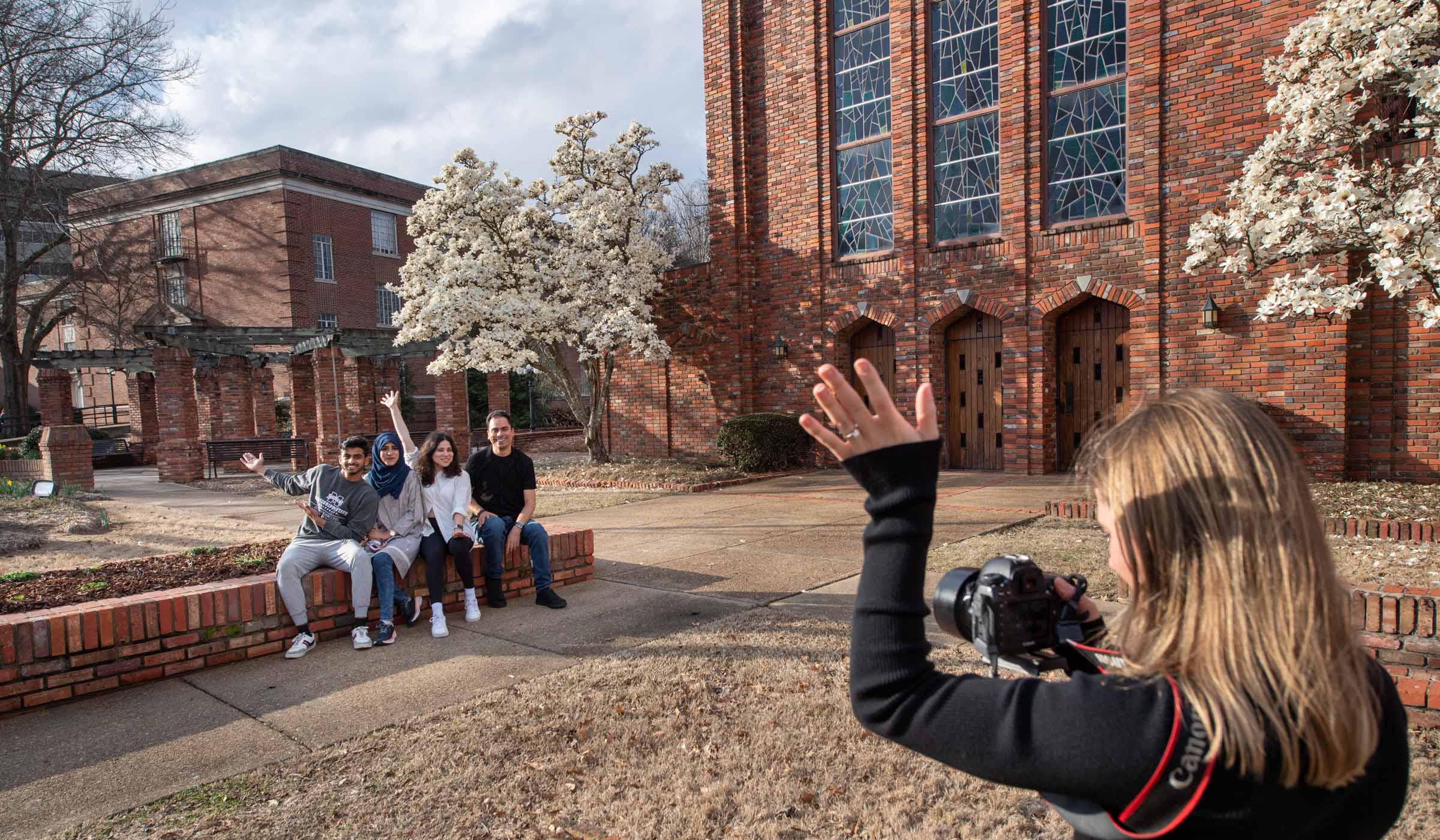 Four international students seated on the brick planter outside the Chapel of Memories wave back at the photographer in the foreground. The Japanese manolia trees in the background are full of white blossom.