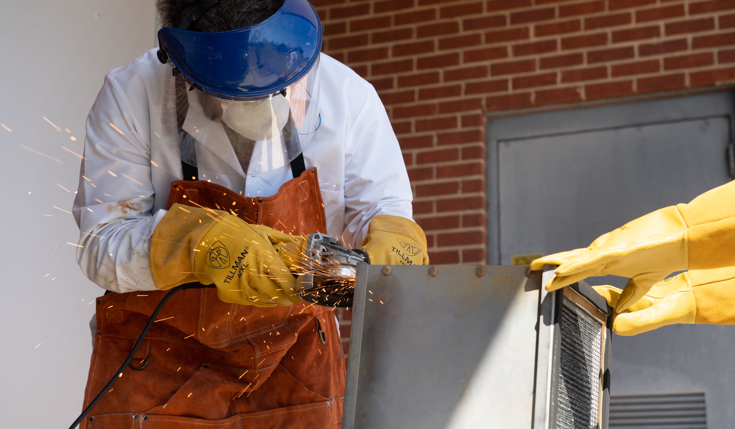 Sparks fly from a saw cutting through the metal casing of an an axial filter. The saw is operated by staff in an apron, wearing a protective apron, yellow gloves, a facemask and eye protection, with a pair of hands in yellow gloves holding the filter stead on the right.