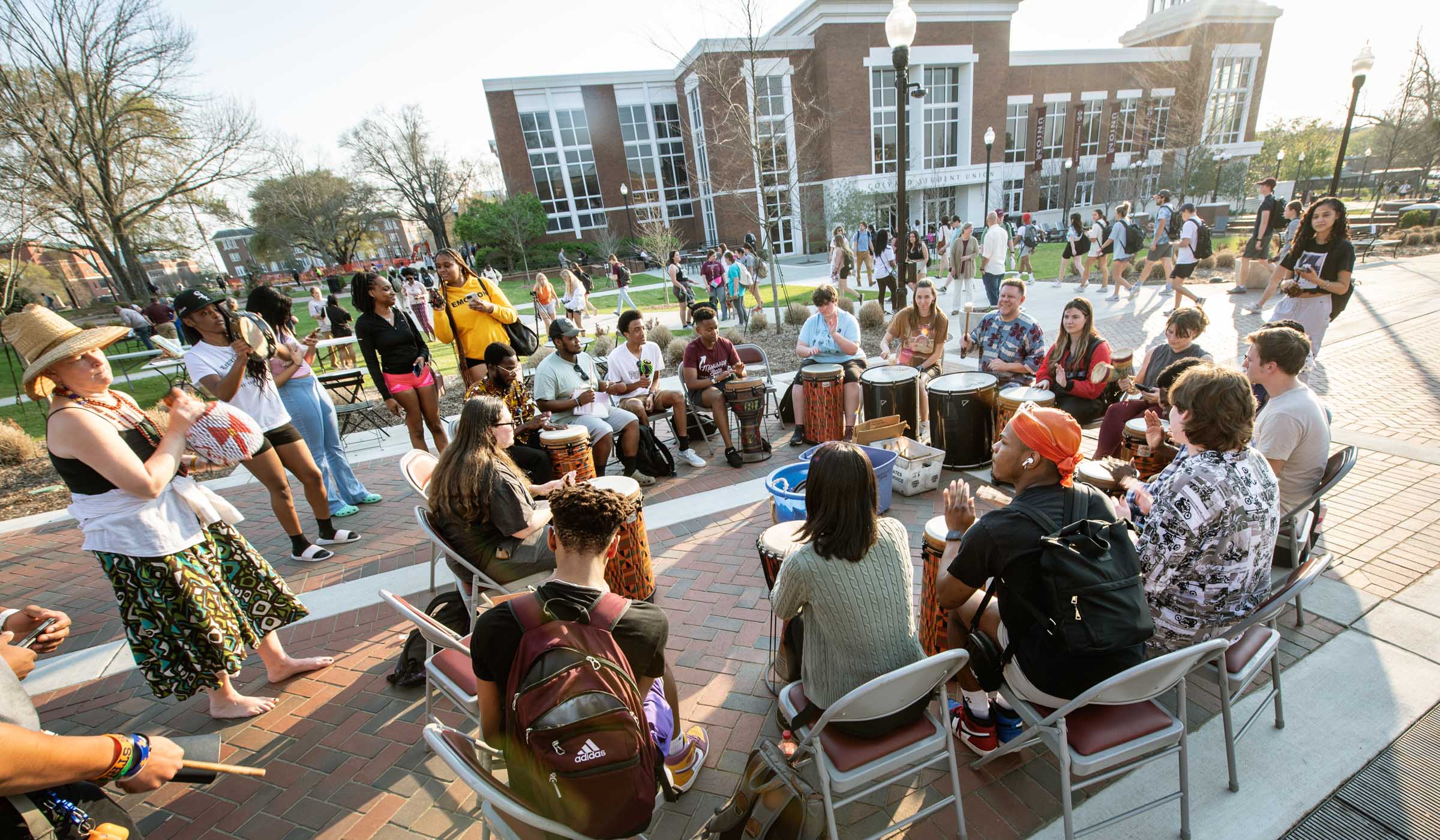 A drum circle plays in the setting sun on the YMCA Plaza, with Colvard Union and passersby in the background.
