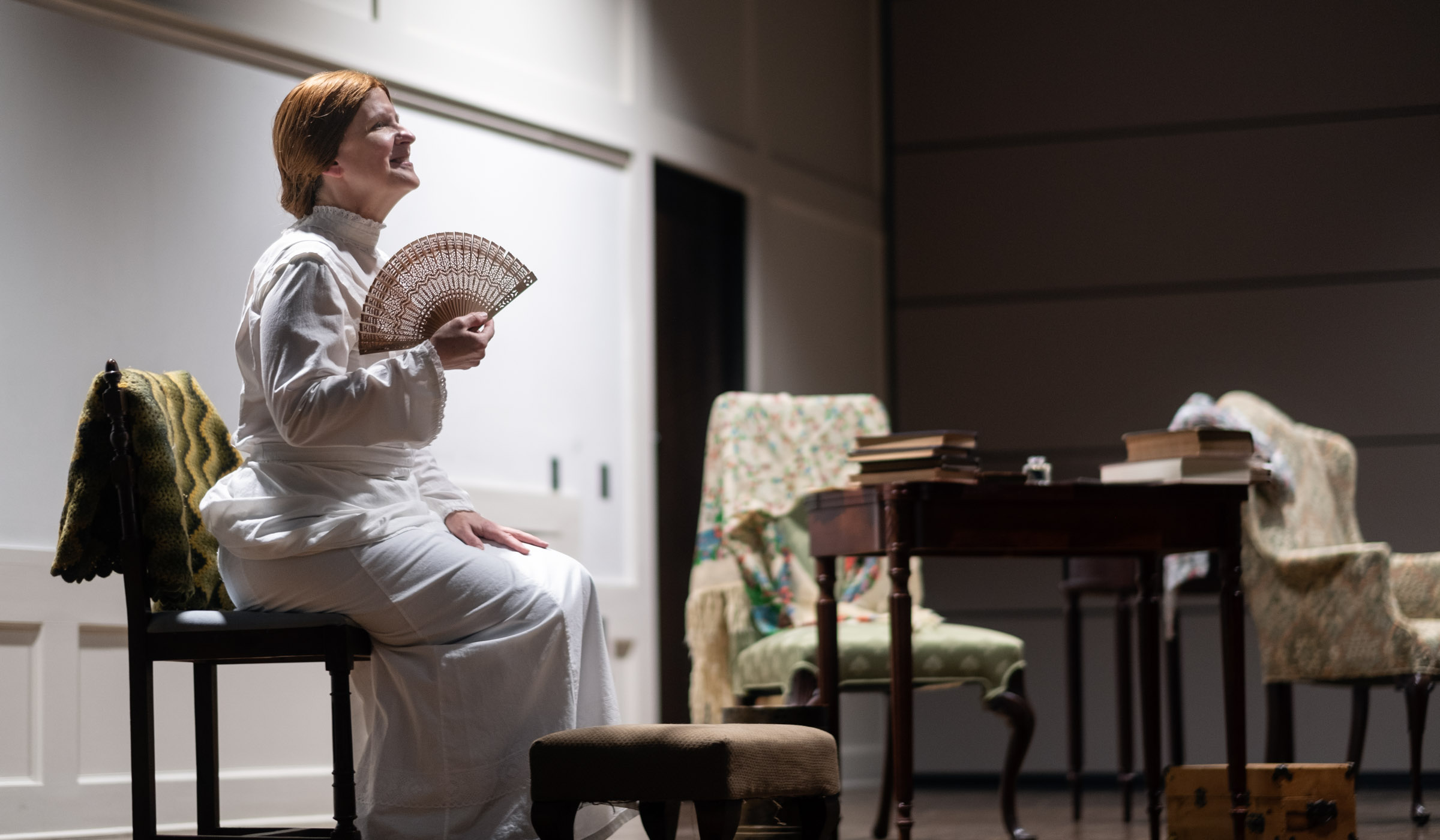 Actress Kennedy Miazza channels Emily Dickinson on the stage, wearing a white period dress and waving a fan while seated is seated on a historic chair.