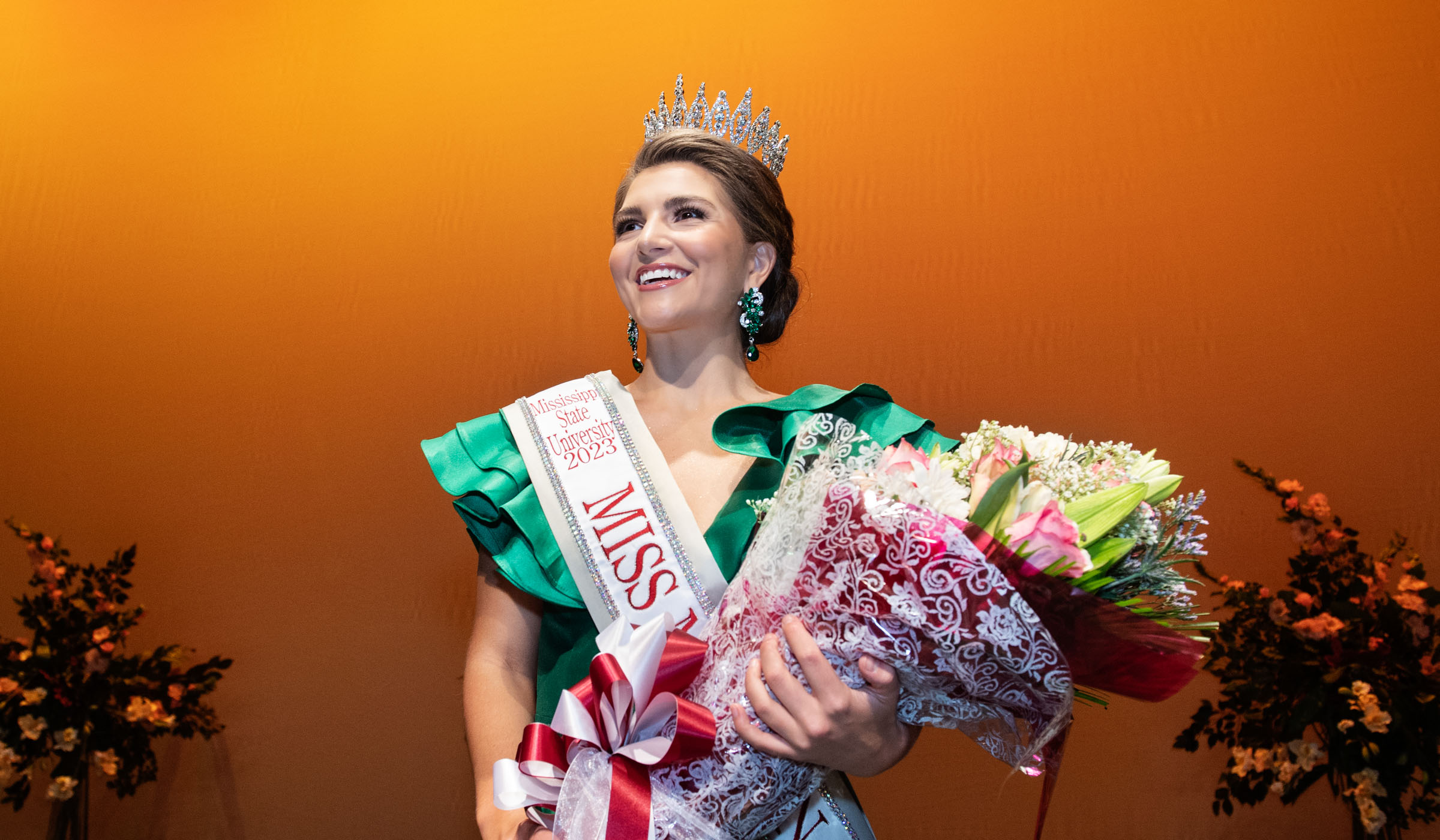 Miss Maroon &amp; White 2023 Madeleine Thompson just after winners were announced: with orange stage backdrop, holding flowers, and wearing Miss Maroon &amp; White sash and crown.