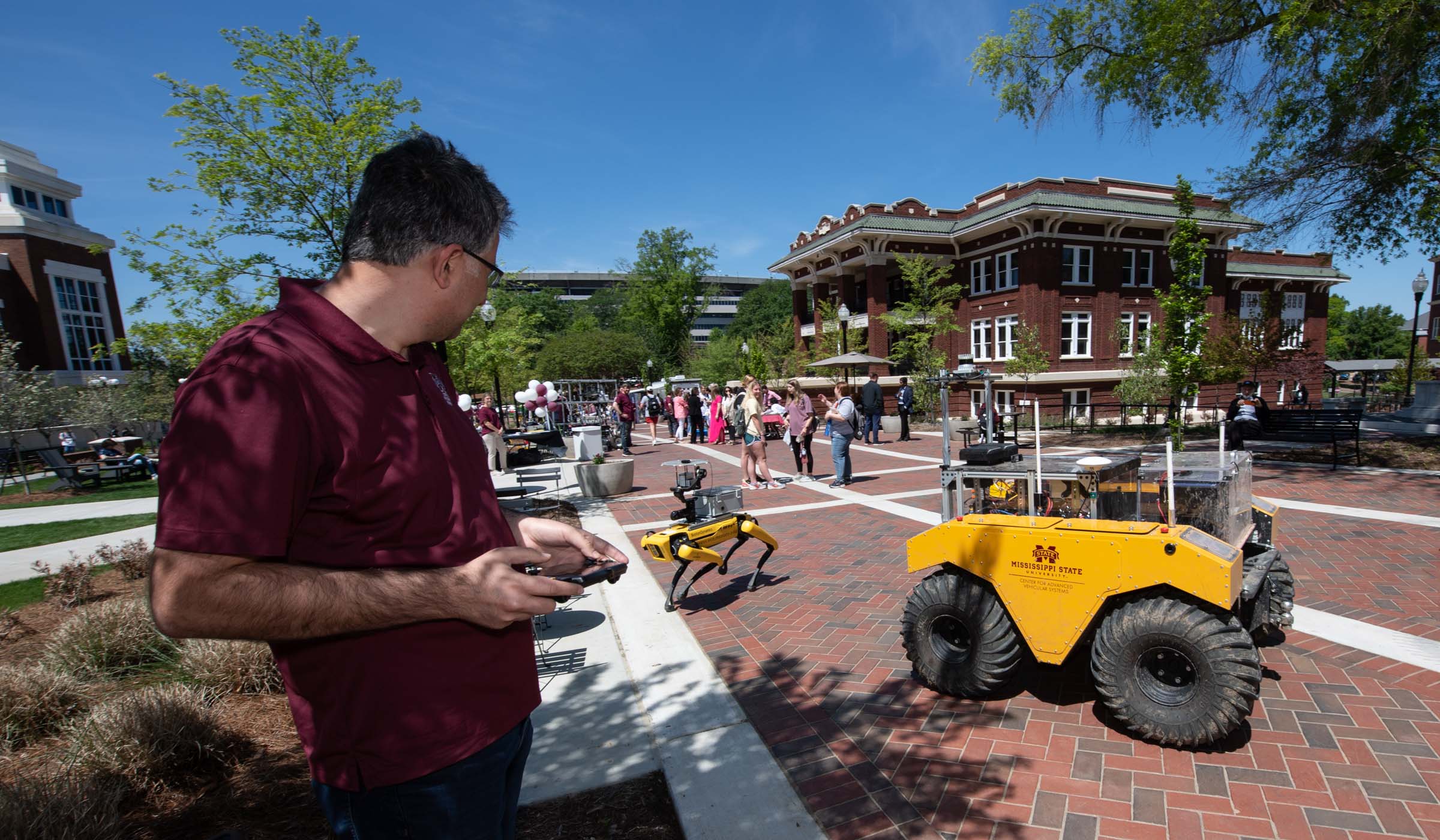 Looking West down the YMCA plaza with research demo booth stretching along the sides, two yellow robots (one with animal-like legs a larger one with wheels) operate in the foreground.