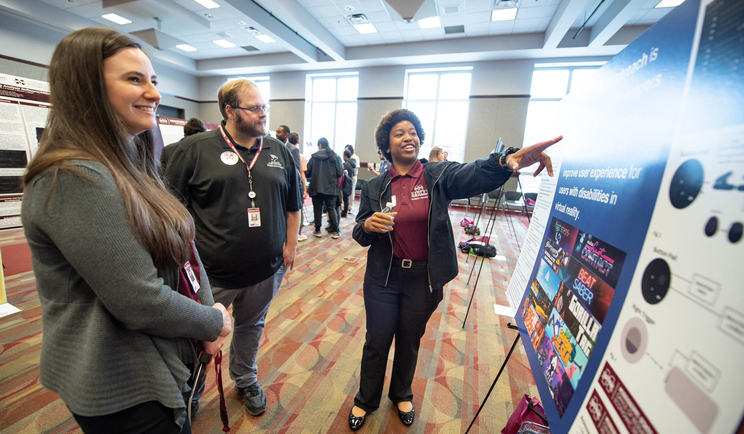 At center, a student points to her poster research, with two onlookers to her left, all backlit with the windows of the Colvard Union ballroom.