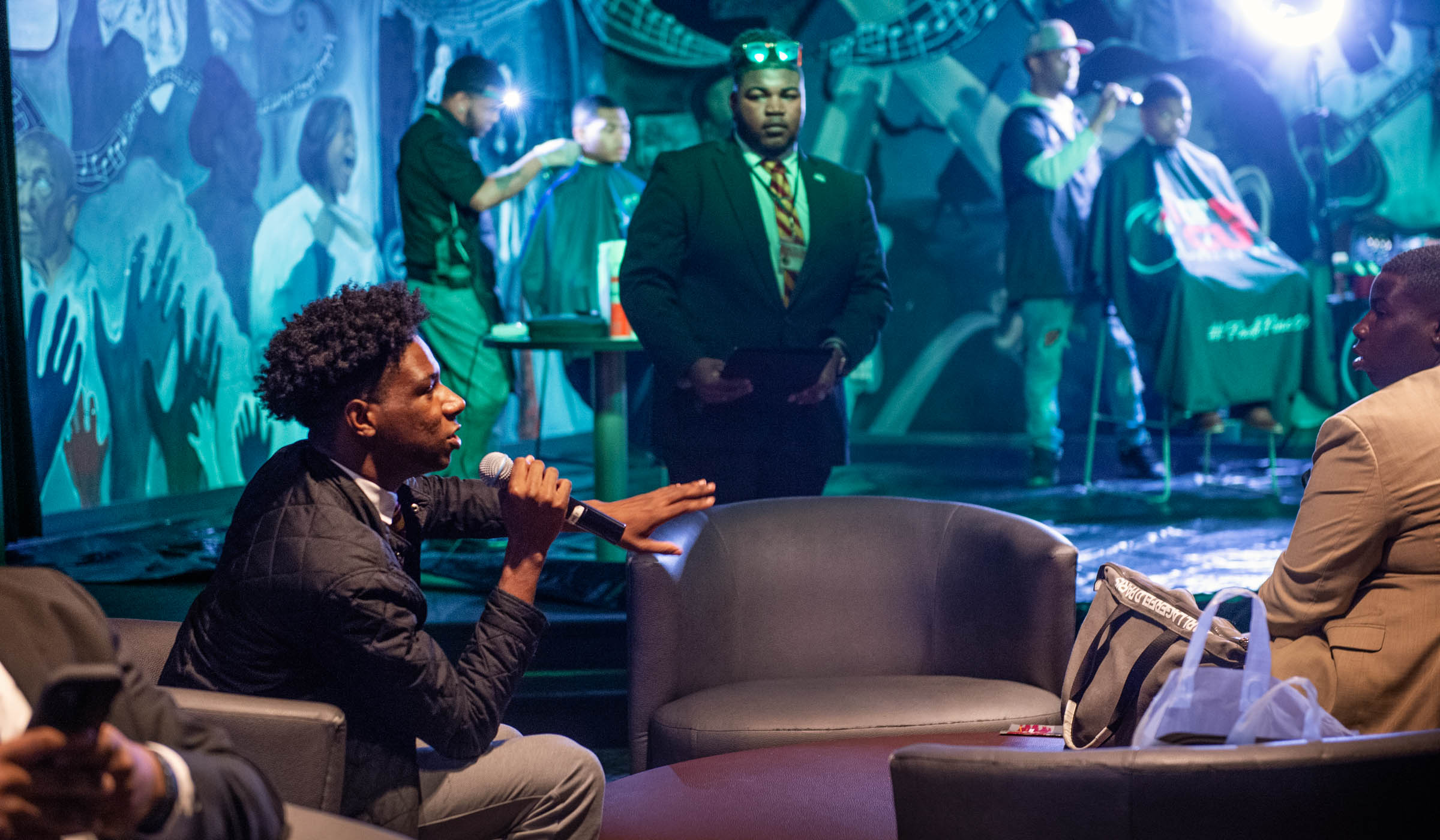 With two barbers giving haircuts to students on the Dawg House stage behind him, student Jaden Joiner speaks into a mic while seated with others in armchairs.