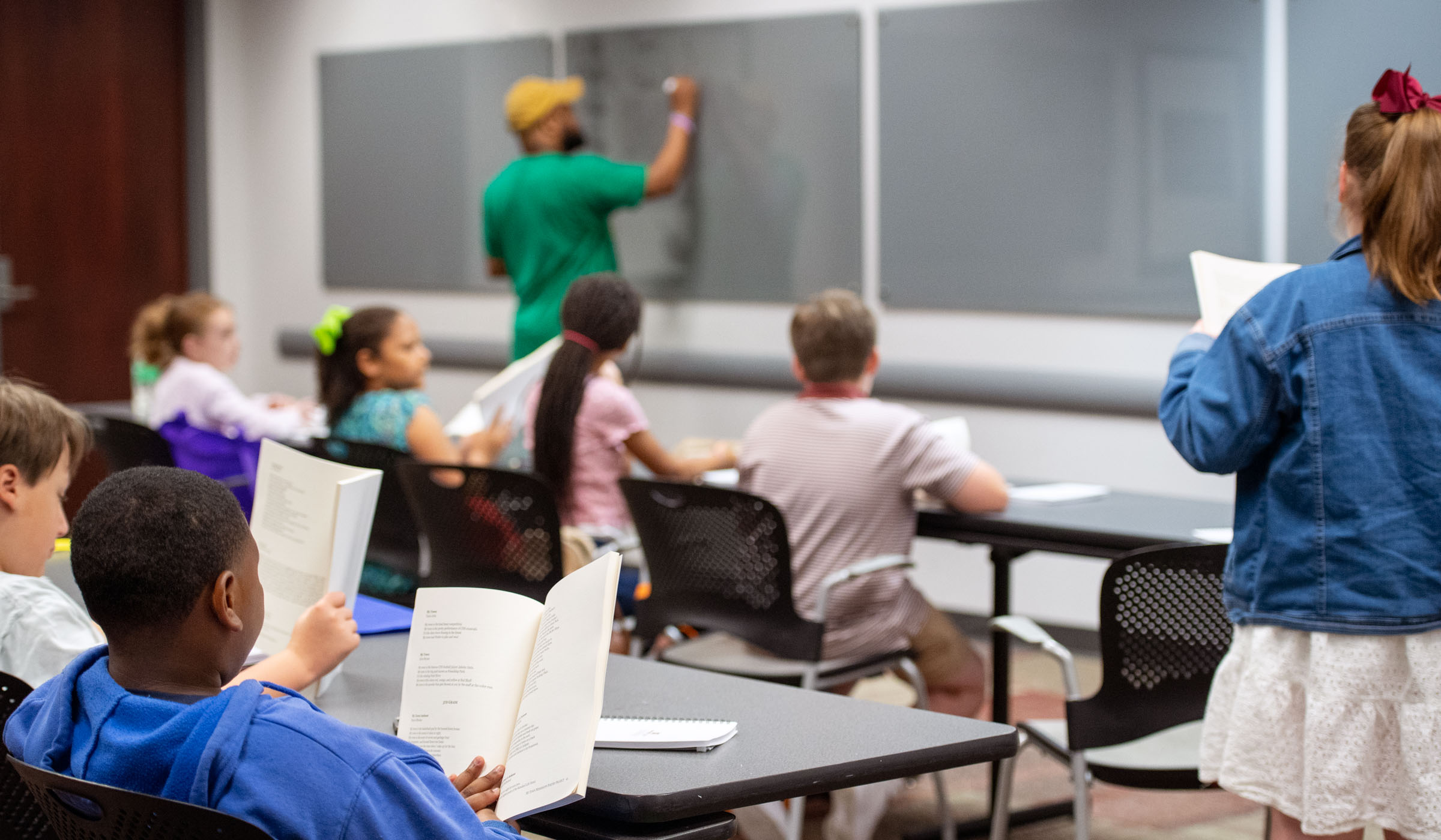 In a classroom filled with children holidng poetry anthologies (photographed from behind), one girl stands at the far right, reading aloud, while a teacher takes notes on a grey writeboard.