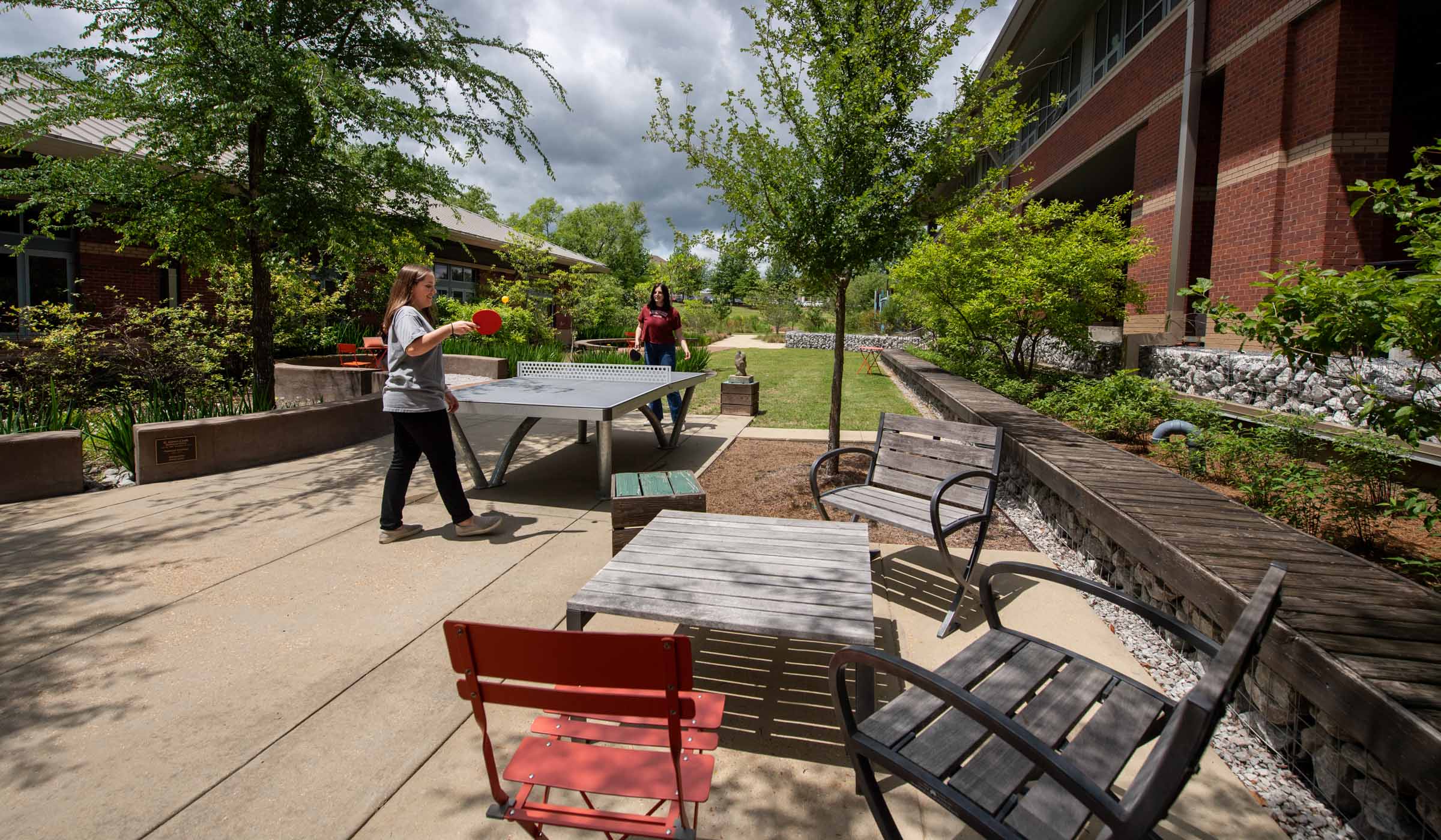 The sunlit Landscape Architecture courtyard featuring chairs and a table in the foreground and two students playing pingpong at center.