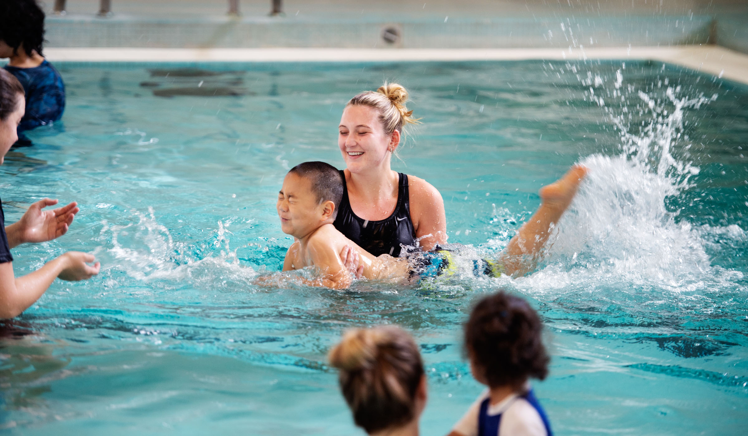 Child Jeffrey Zhang splashily kicks towards an applauding iCanSwim staff member in Sanderson Pool, with assistance of MSU Student Whitney Strong.