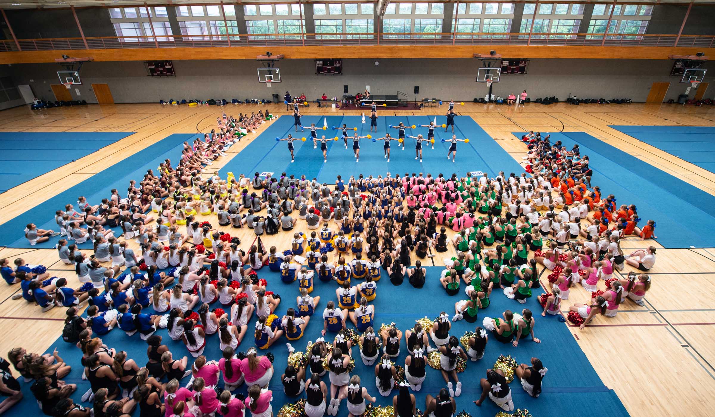 Looking down onto Sanderson&#039;s Main Gym, with blue mats covering the floor and hundreds of cheerleader teams (seated with backs to camera) watching their counselors do a cheer routine.