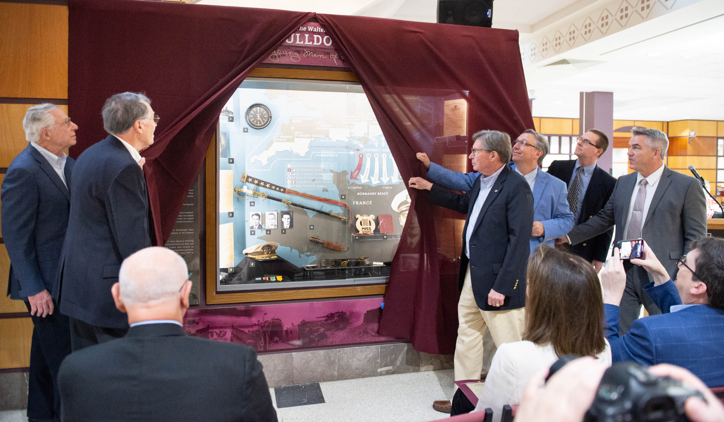 Dedication event participants pull back the maroon curtain, revealing the lit glass case containing WWII alumni Wallace&#039;s artifacts and memorbilia.
