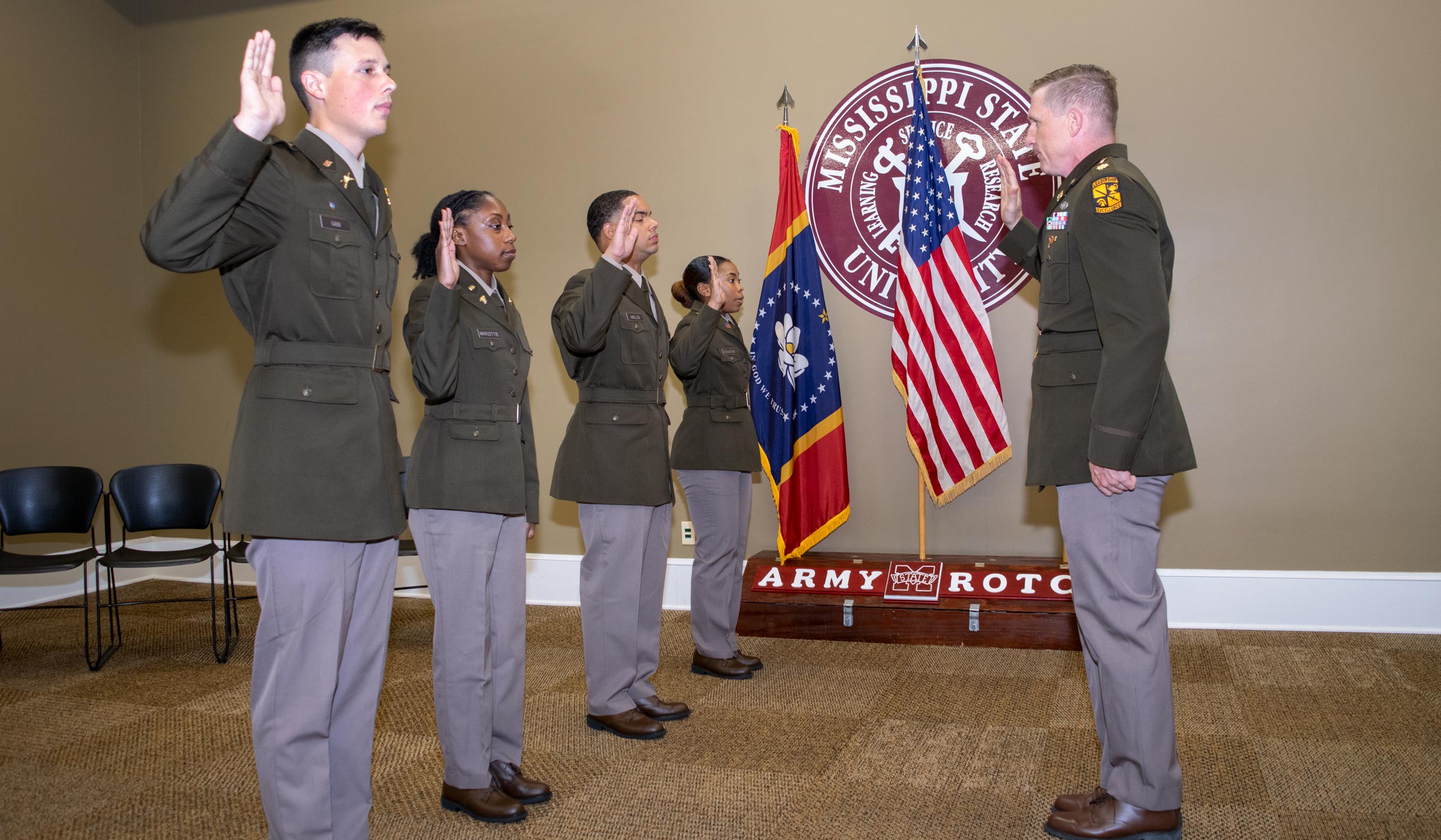 Individuals in green army dress uniforms taking oath in front of American and state flags