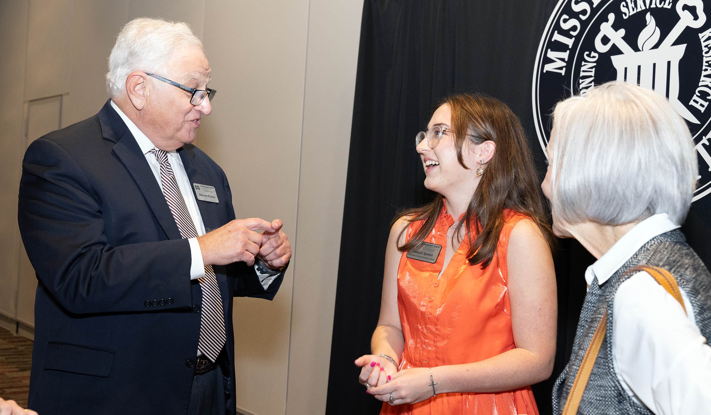 Older male in suit speaking to college age female in orange dress and older female in gray vest and white shirt