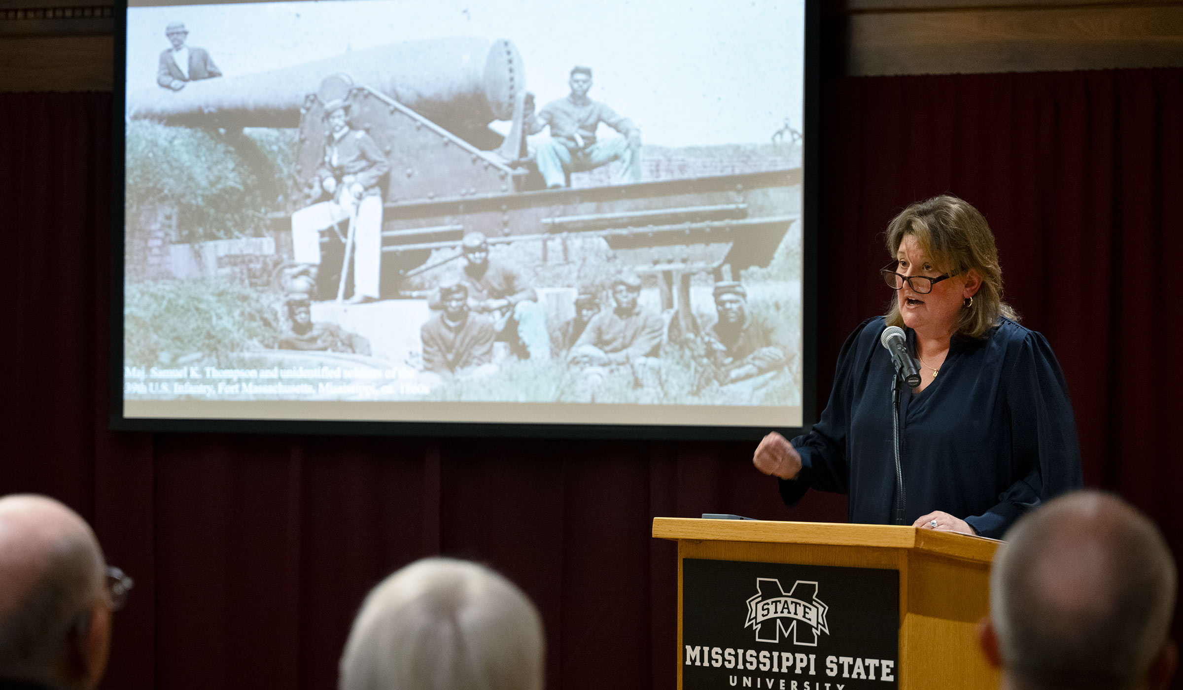 Woman at podium speaking to seated group about the image in background.