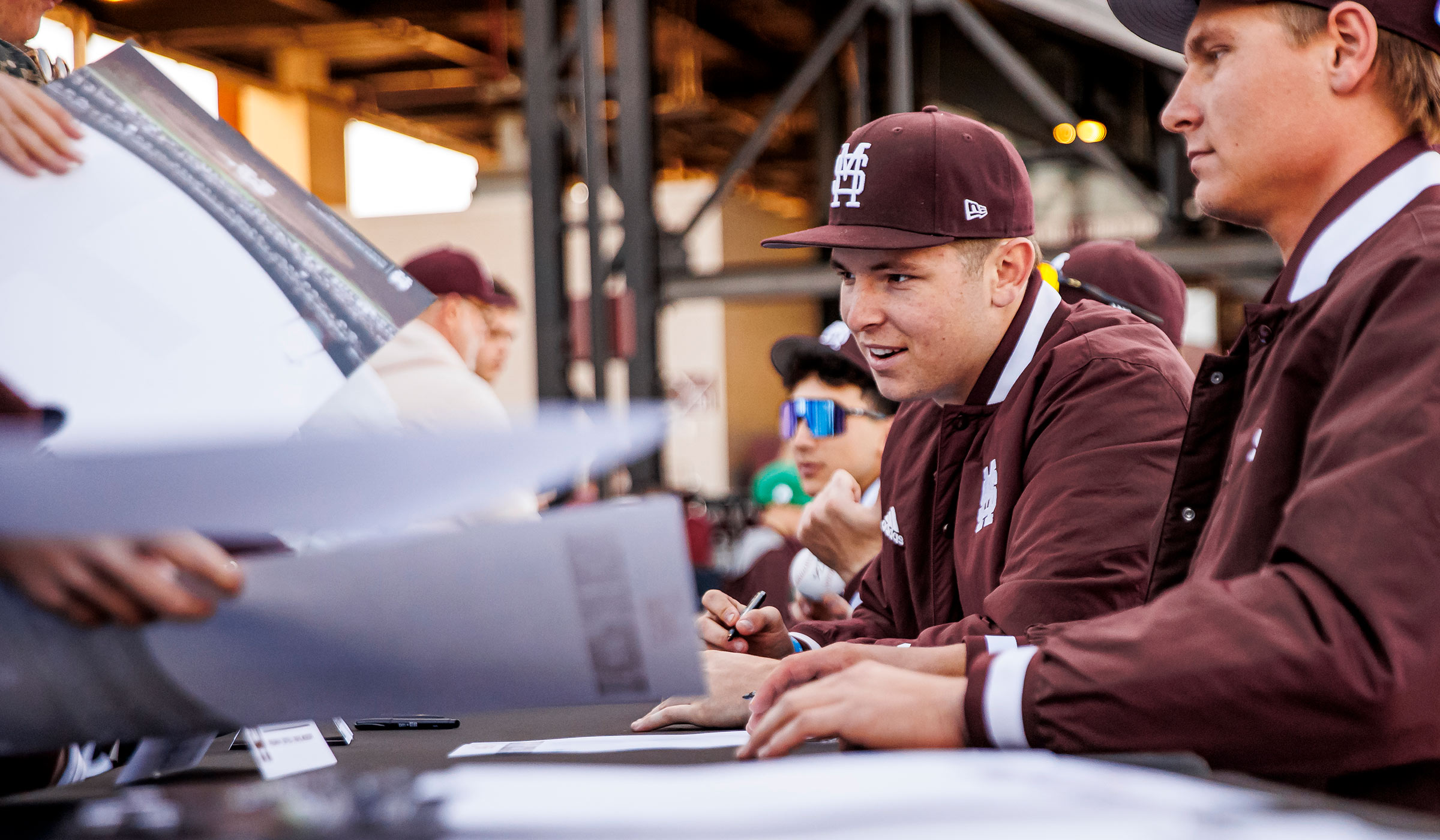 Guy in maroon jacket and baseball cap with pen signing posters