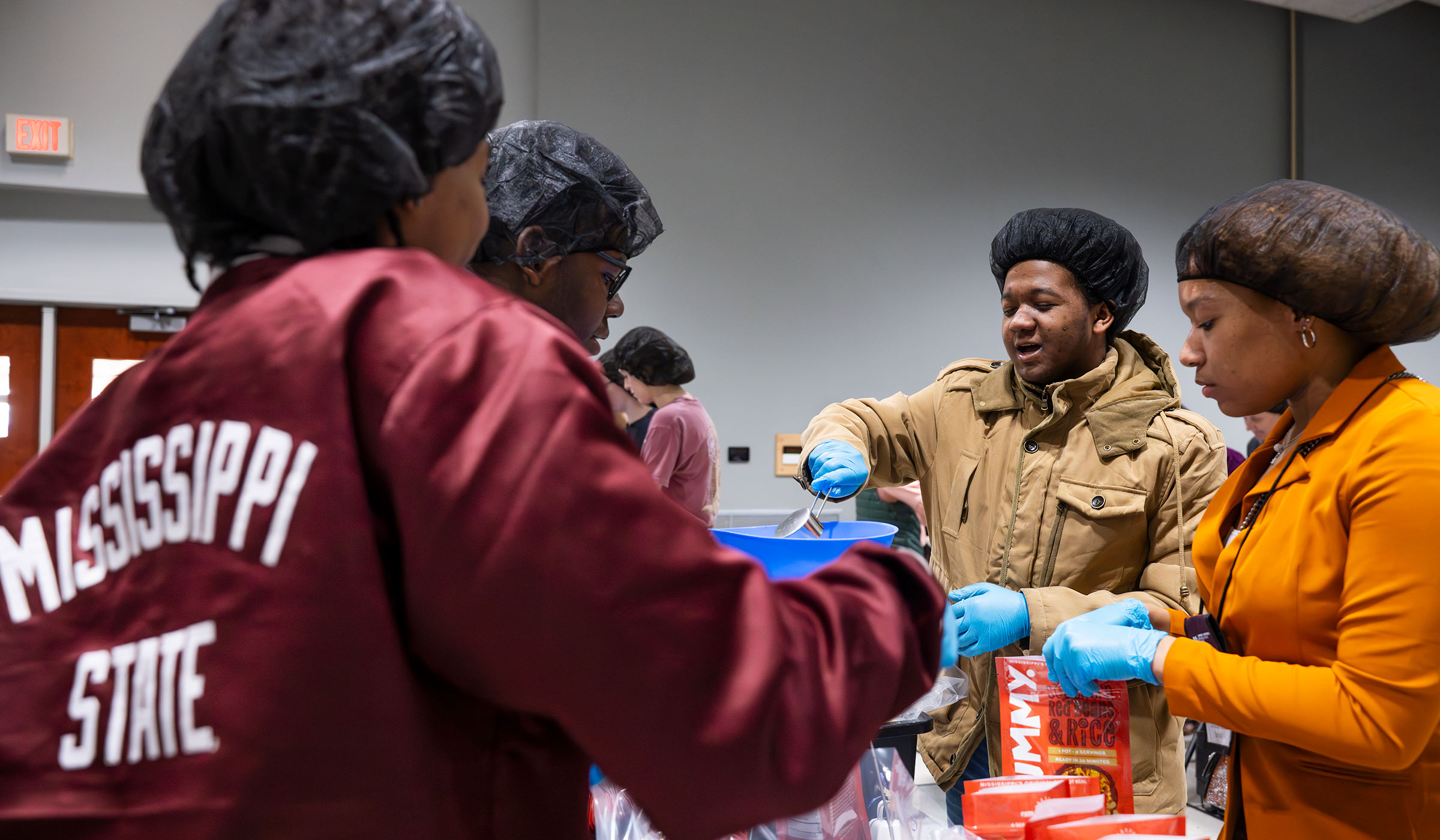 Students in brown, orange, and maroon jackets with hair nets and gloves adding scoops of beans and rice to orange package.