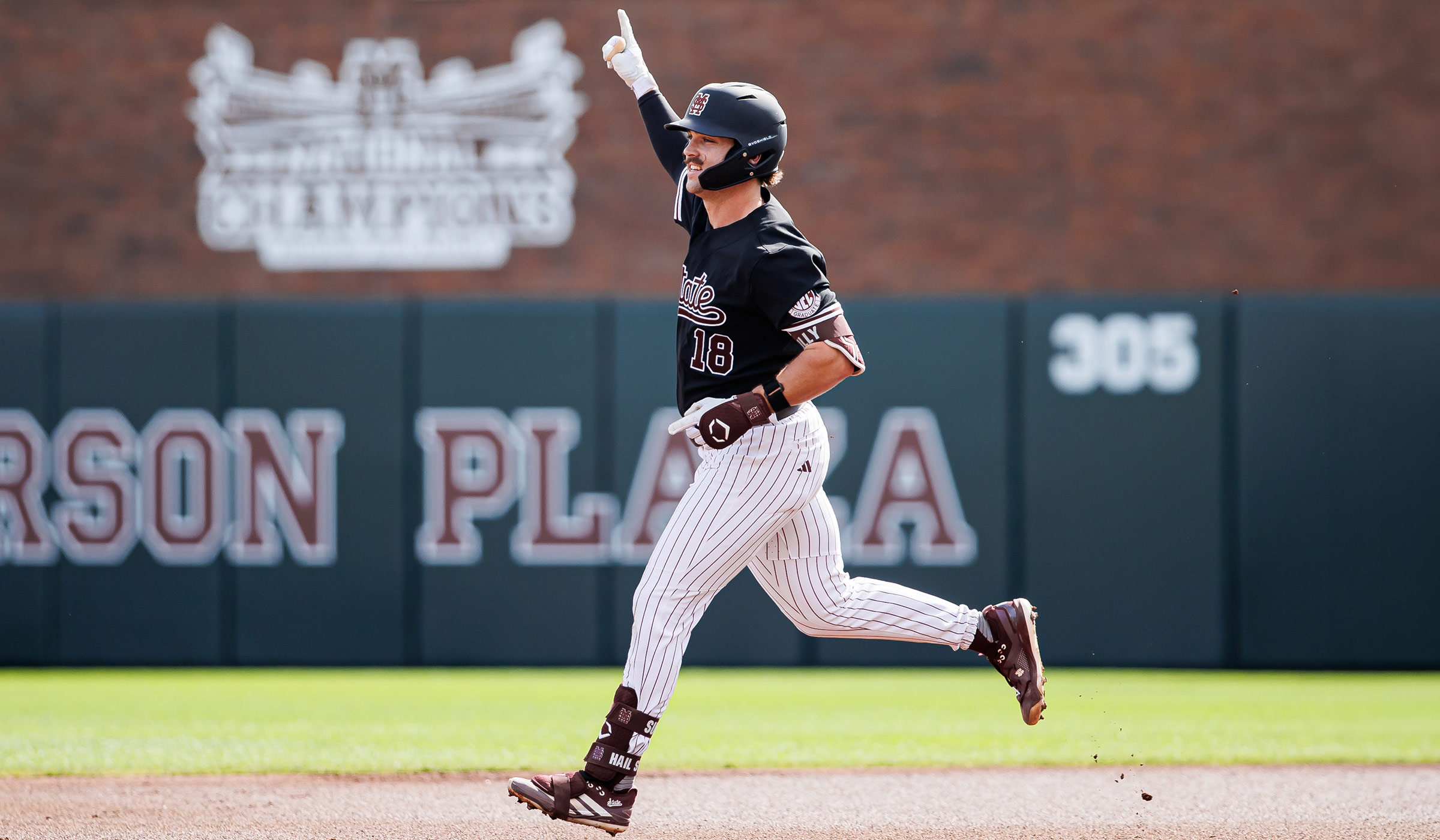 Man in baseball uniform of black helmet, black jersey with maroon accents and white pinstripe pants running bases with hand raised in number one signal.