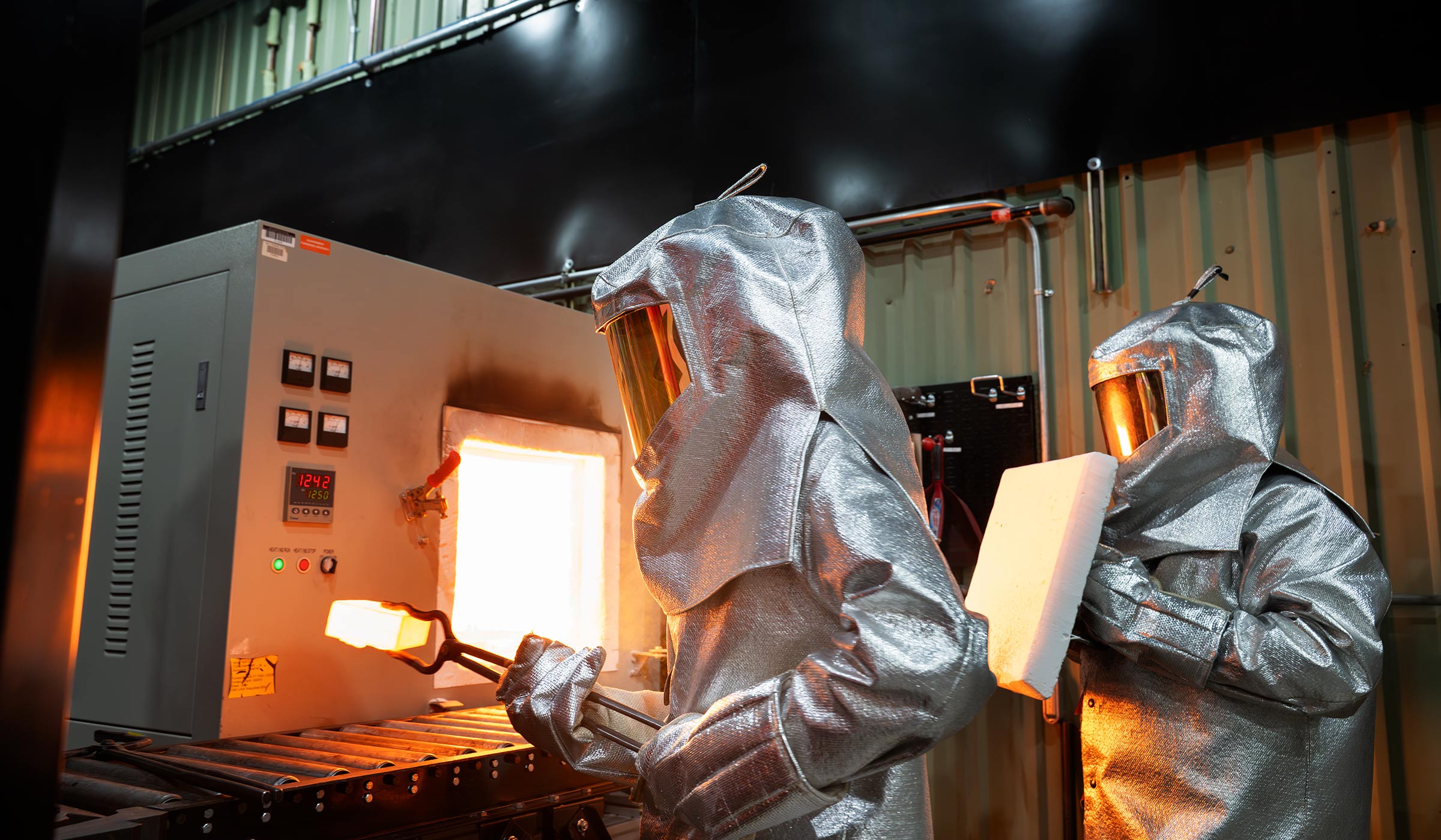 Giant silver box with glowing orange light emanating towards to people dressed in full silver heat-resistant head-to-toe suits with one carrying a white glowing block with giant tongs.