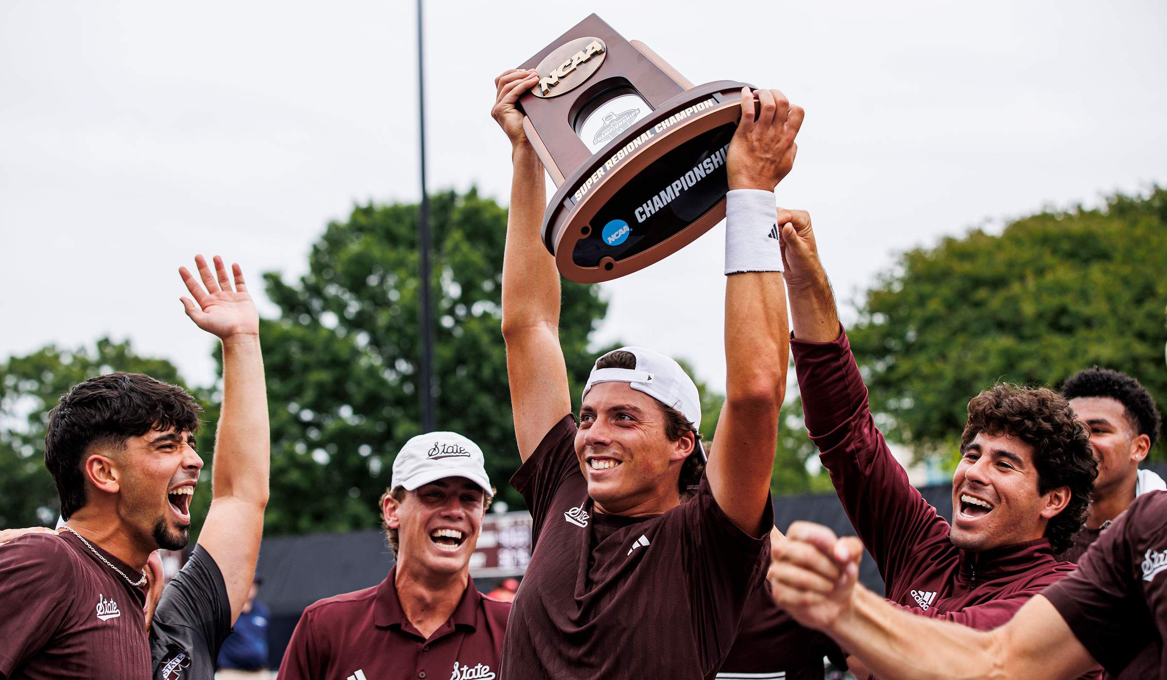 Men in maroon shirts laughing and celebrating while surrounding one guy that is holding up NCAA Super Regional Champions trophy above his head