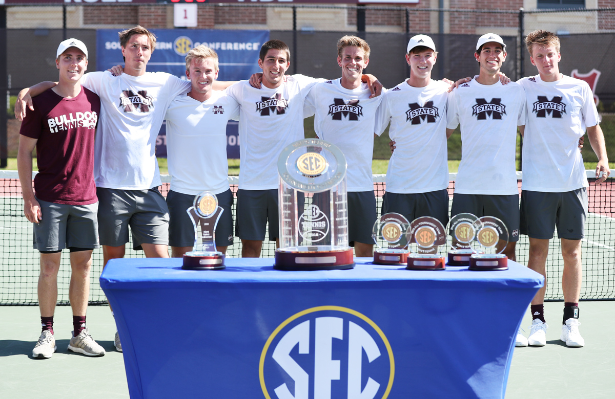 Group photo of eight Men&amp;#039;s Tennis team members standing behind SEC Tennis trophies on a table at the courts.
