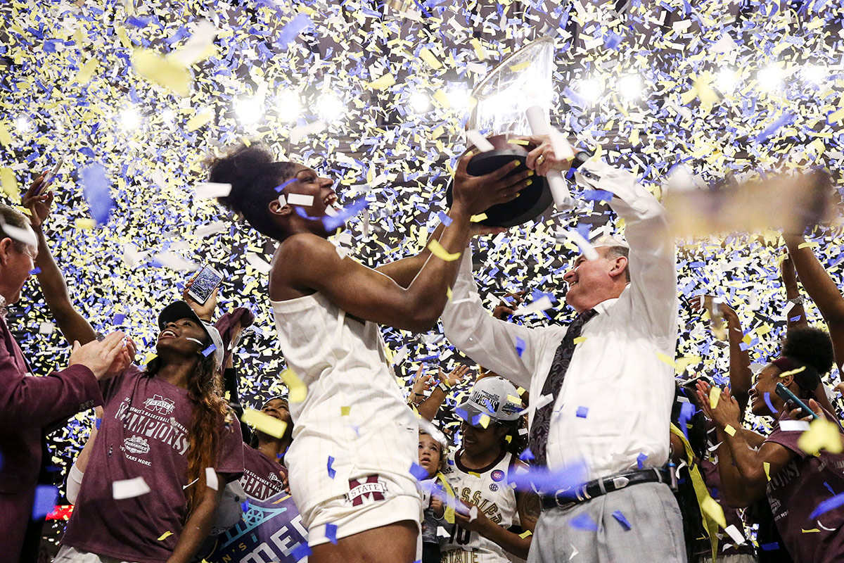 Tall female basketball player and coach in tie holding up trophy with blue and yellow confetti falling around them