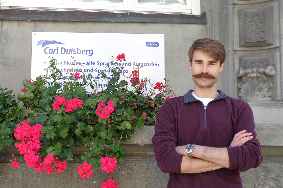 Ryan Shoemake stands near a bush of bright pink flowers in front of the Carl Duisberg Centren in Radolfzell, Germany.