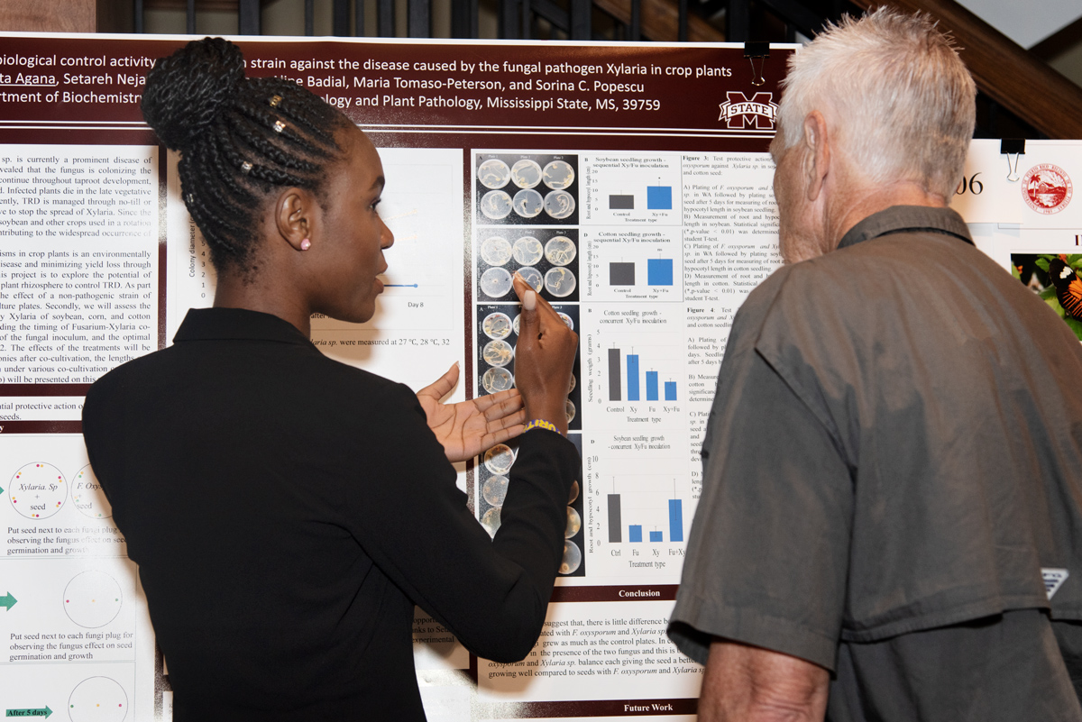 A student gestures at a large research board to explain her research project to a spectator at the symposium.