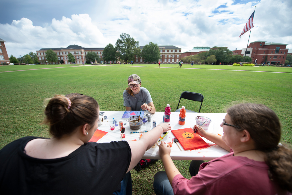 Three female students share a laugh while paint canvases at a table with the Drill Field behind them.