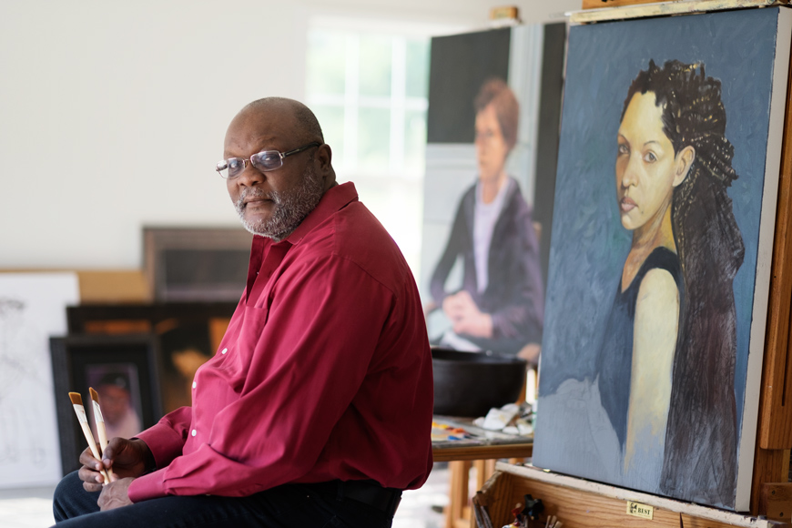 Alex Bostic, with paint brushes in hand, sits in front of a canvas depicting a woman with long dark hair.