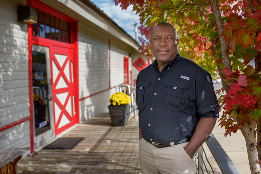 Harvin Hudson in a blue shirt and khaki pants standing outside in front of a building with a red door.