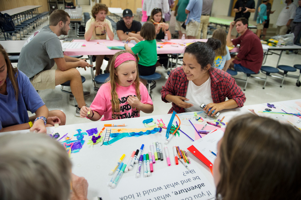 MSU students and children brainstorm at a school cafeteria table covered with drawing paper and art supplies.
