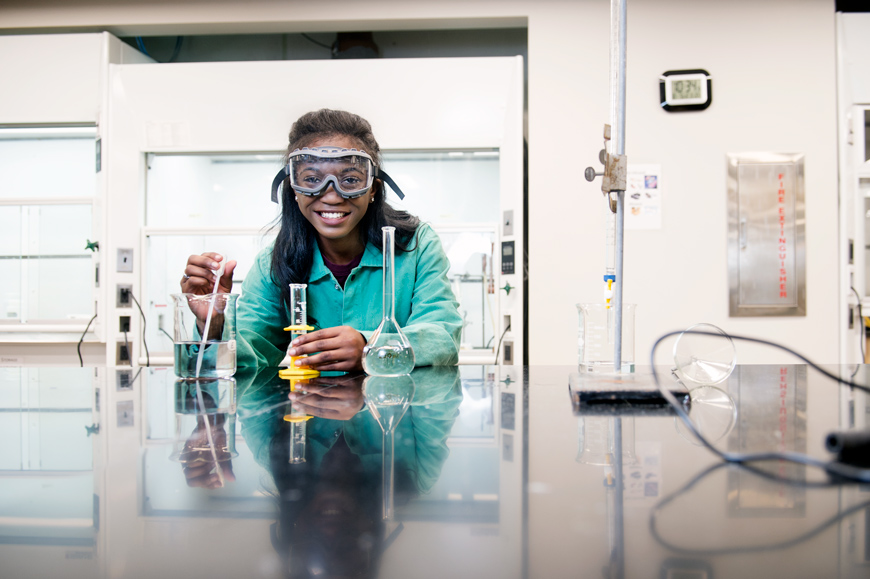 Dajaina Martin dressed in a green over coat and safety glasses in a lab as she works on her degree and major in biochemistry.