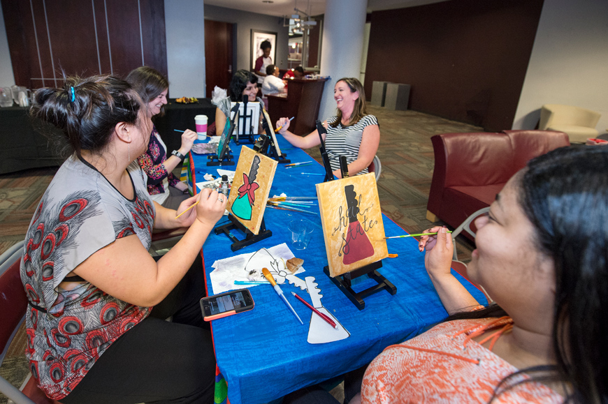 Female MSU Employees sit at table in The Dawg House, making paintings of cowbells.