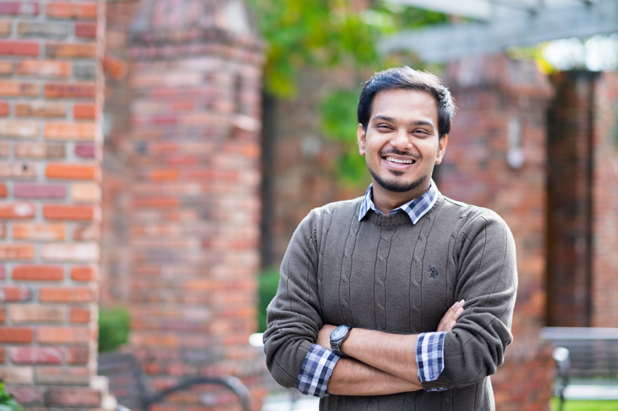 Shuvam Saha, Mississippi State University student, wearing a grey sweater and plaid shirt while smiling and standing in front of brick and wood arbors and benches near the MSU Chapel of Memories