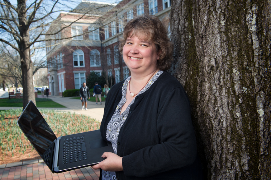 Tamra Swann leans against a tree on the Drill Field while holding a computer.