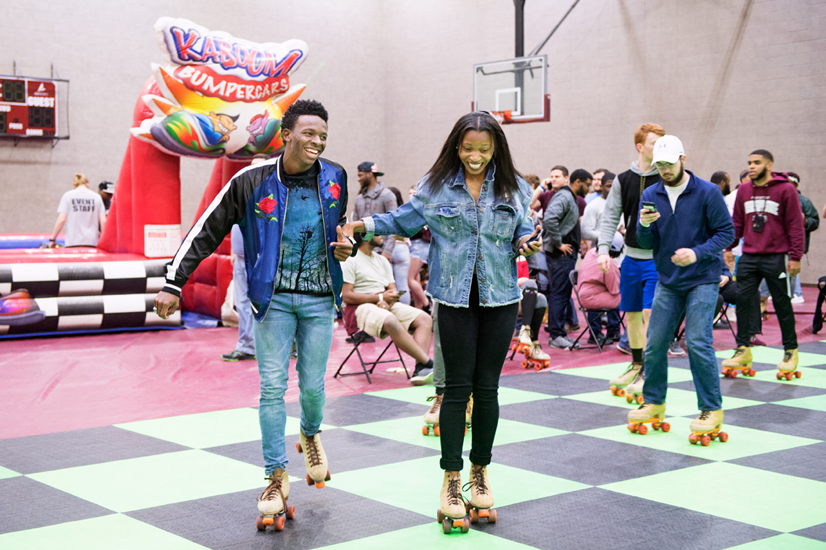 Two students rollerskating on checkerboard floor with bumper cars in background