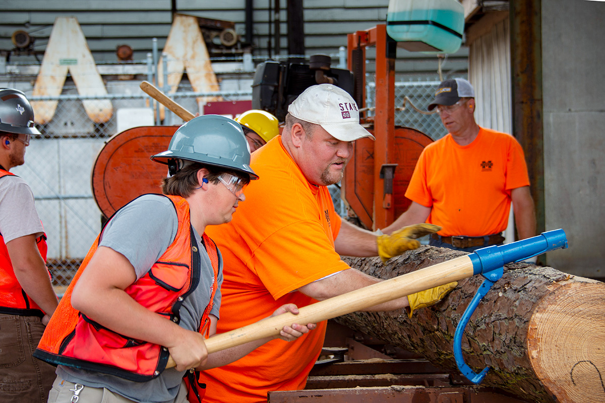 Man in orange shirt and male camper in orange vest and gray helmet using a blue hook to aid in sawing logs