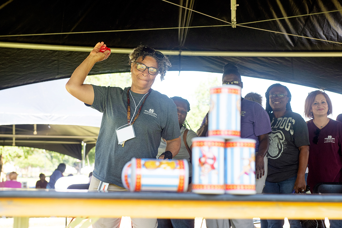 Woman in gray shirt about to throw red bean bag at plastic bottles in carnival style game