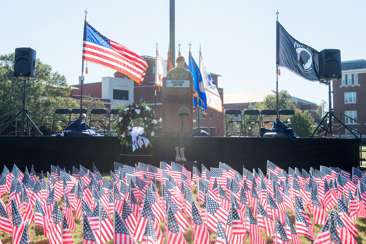 Cadet in Army uniform reading names of the fallen between United States flag and POW flag.