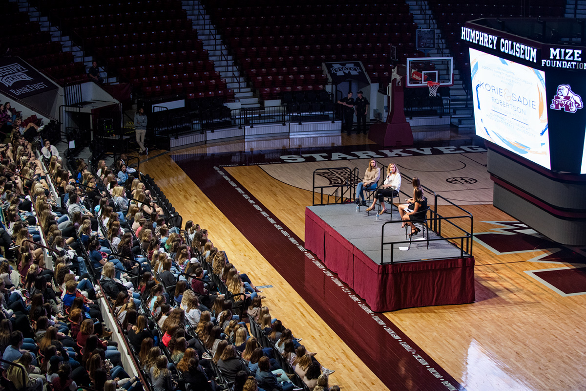 Two students interviewing two female guests on stage in Humphrey Coliseum in front of crowd.