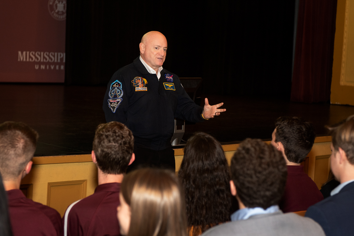 Man in dark blue jacket with astronaut patches speaking to students