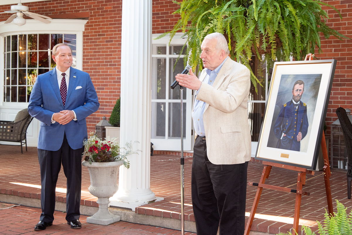 Man in light khaki blazer speaking at microphone near painting of Ulysses S. Grant with man in checkered blazer in background.