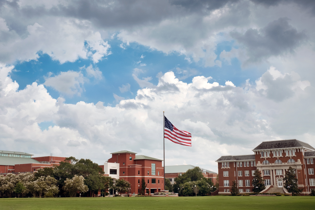 Summer clouds build and blow the American flag over a wide view of the Drill Field toward Mitchell Memorial Library.