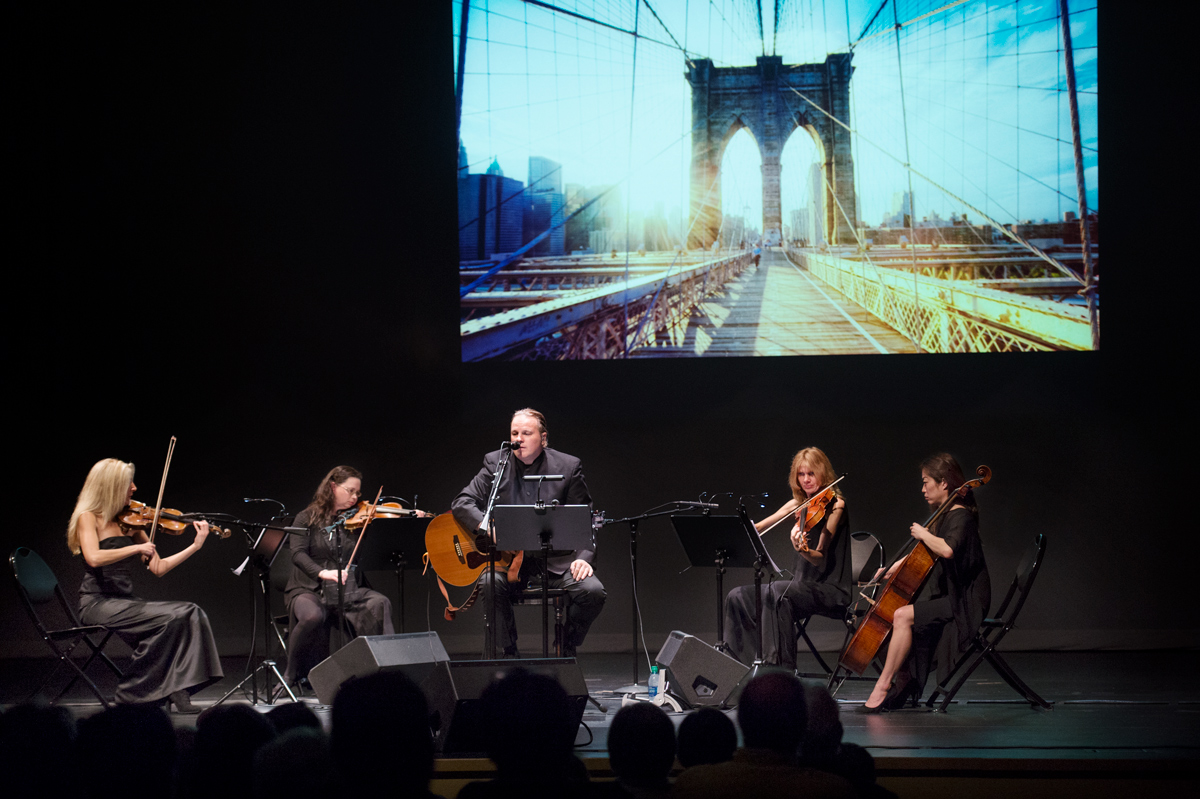 A string quartet surrounds the Cellophane Flowers lead singer on guitar, with a photo of the Brooklyn Bridge projected behind.