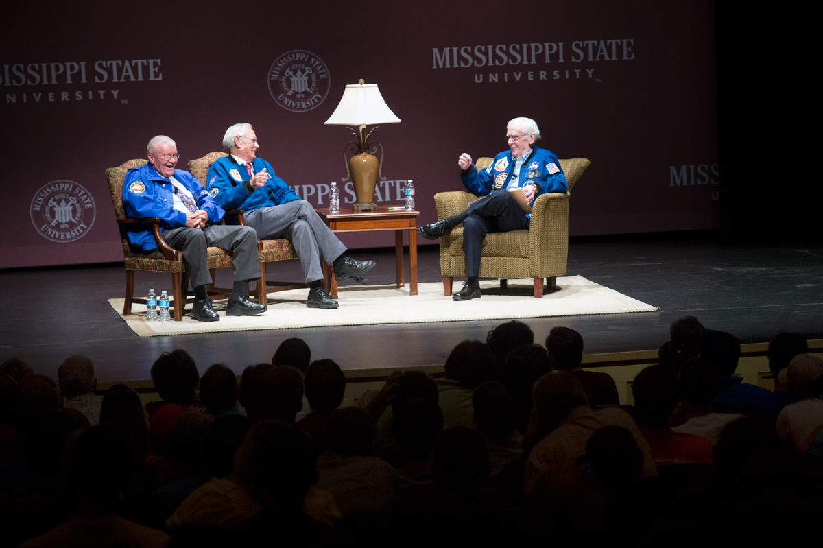 Seated in a living room set on the stage of Bettersworth Auditorium, Apollo astronauts Charlie Duke and Fred Haise share a laugh