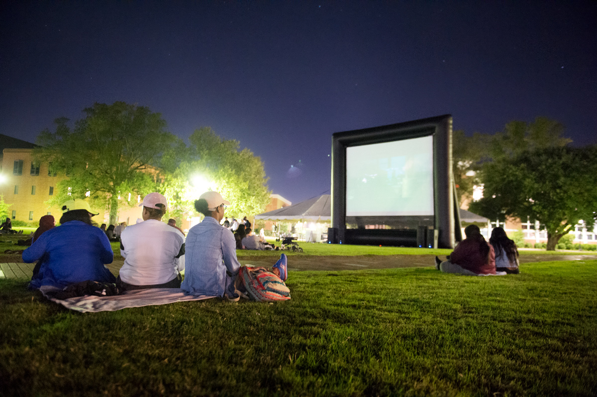 Students sit on blankets on the Drill Field grass at night, while the movie screen glows in the background.